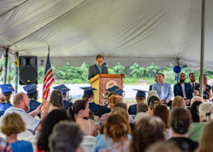 Man wearing a suit standing behind a wooden podium giving a speech to graduates and audience members